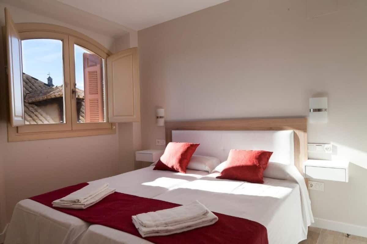 Un lit double avec une literie rouge et blanche, une tête de lit en bois et une fenêtre donnant sur des immeubles.