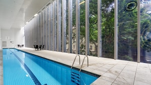 An indoor swimming pool with clear blue tiles, a metal ladder, and a view of trees through large windows. - Bright 1-bed Retreat in Charlotte Towers (Brisbane)