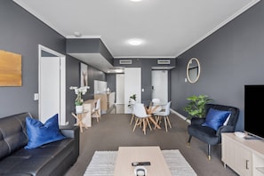 Ein modernes Wohnzimmer mit Sofa, Couchtisch und Fernseher. Es gibt einen Essbereich mit Tisch und Stühlen. Die Wände sind grau und es gibt eine Deckenleuchte.