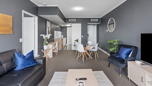 A modern living room with a sofa, coffee table, and a TV. There is a dining area with a table and chairs. The room has grey walls and a ceiling light. - Bright 1-bed Retreat in Charlotte Towers (Brisbane)