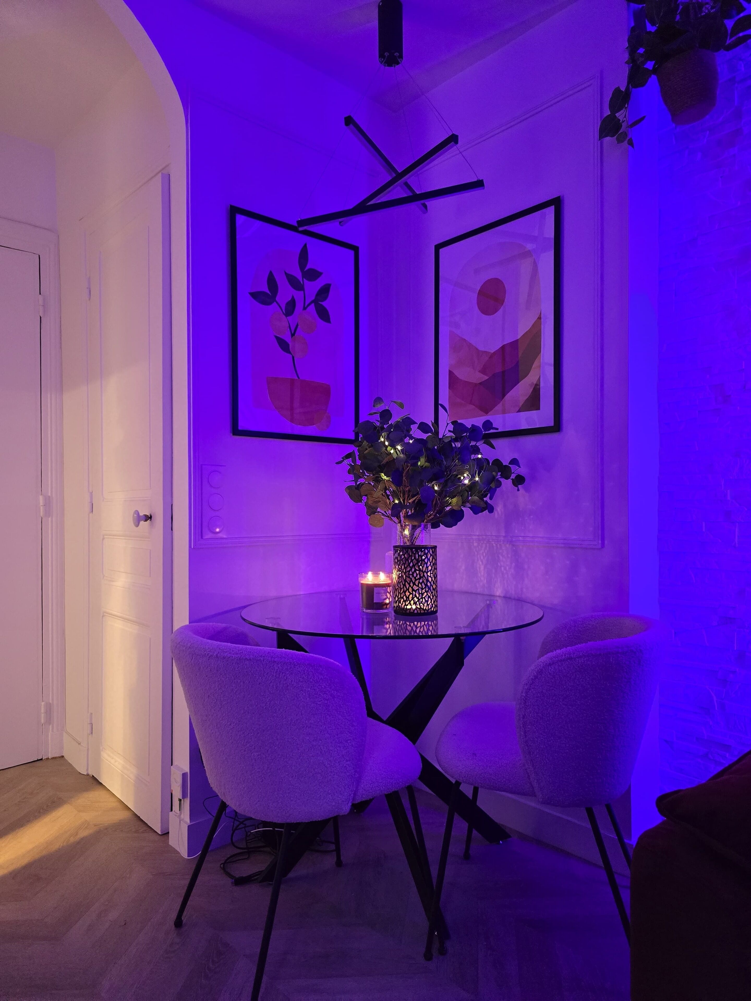 Un comedor con una mesa de vidrio, dos sillas, un jarrón con flores y cuadros en la pared.