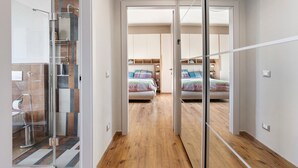 Moderni hotellihuone, jossa on lasiseinäinen suihkutila, puulattia ja näkymä avoimesta ovesta toiseen huoneeseen, jossa on sänky ja työpöytä.