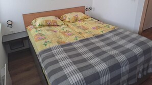 En säng med ett blommigt överkast och en rutig pläd, ett sängbord och en väggmonterad ljusströmbrytare.