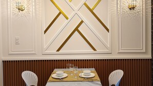 Un coin repas avec une table blanche dressée pour deux personnes, entourée de chaises blanches, adossé à un mur orné de panneaux décoratifs et de luminaires muraux.
