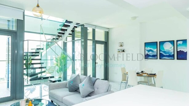A modern living room with a glass staircase, a sofa with cushions, a dining table, and framed artwork on the wall. - Bloomfields 2BR Duplex Canal View Ra (Abu Dhabi)