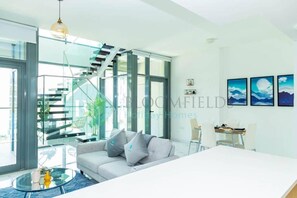 Ein modernes Wohnzimmer mit einer Glastreppe, einer Couch mit Kissen, einem Esstisch und gerahmten Kunstwerken an der Wand.