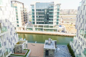 A modern high-rise building with a river in the foreground, a view of the cityscape in the background, and a construction site to the right. - Bloomfields 2BR Duplex Canal View Ra (Abu Dhabi)