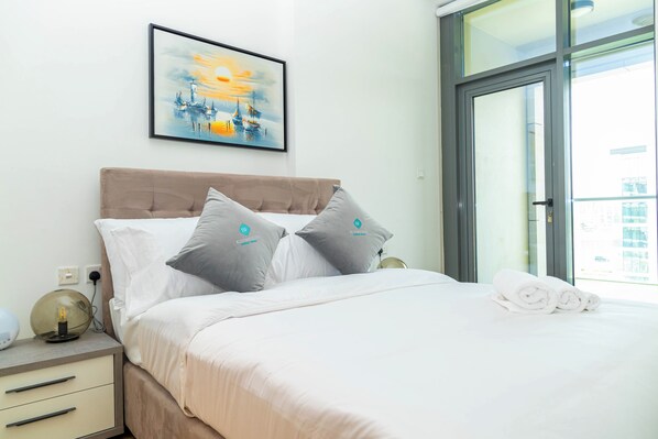A hotel room with a bed, pillows, a nightstand, a lamp, a framed artwork, and a view through a glass door. - Bloomfields 2BR Duplex in Raha Lofts - Raha (Abu Dhabi)