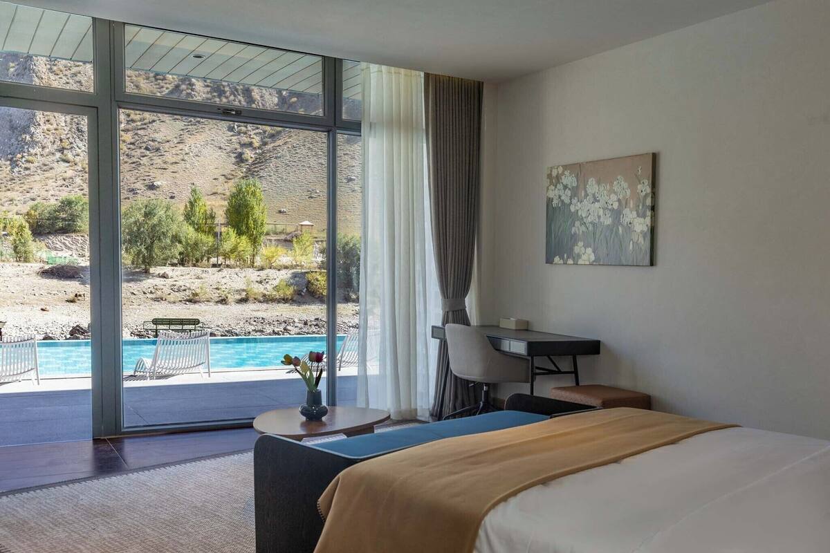 Et hotelværelse med stort vindue, seng, skrivebord og udsigt til pool og bjerge.