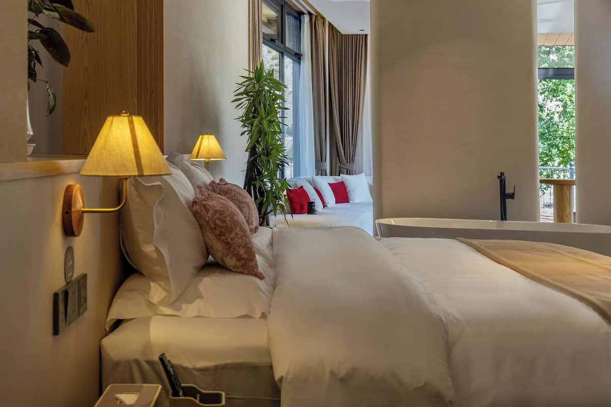 Et moderne hotelværelse med en stor seng, to sengeborde med lamper, en plante og udsigt til det fri.
