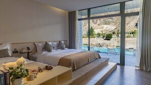 Un dormitorio con una cama grande, una mesita de noche con libros y una planta, y vistas a una piscina y montañas a través de ventanas amplias.