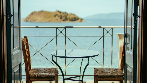 Un balcon ouvert donnant sur une mer calme et une île lointaine, avec deux fauteuils en osier et une petite table.