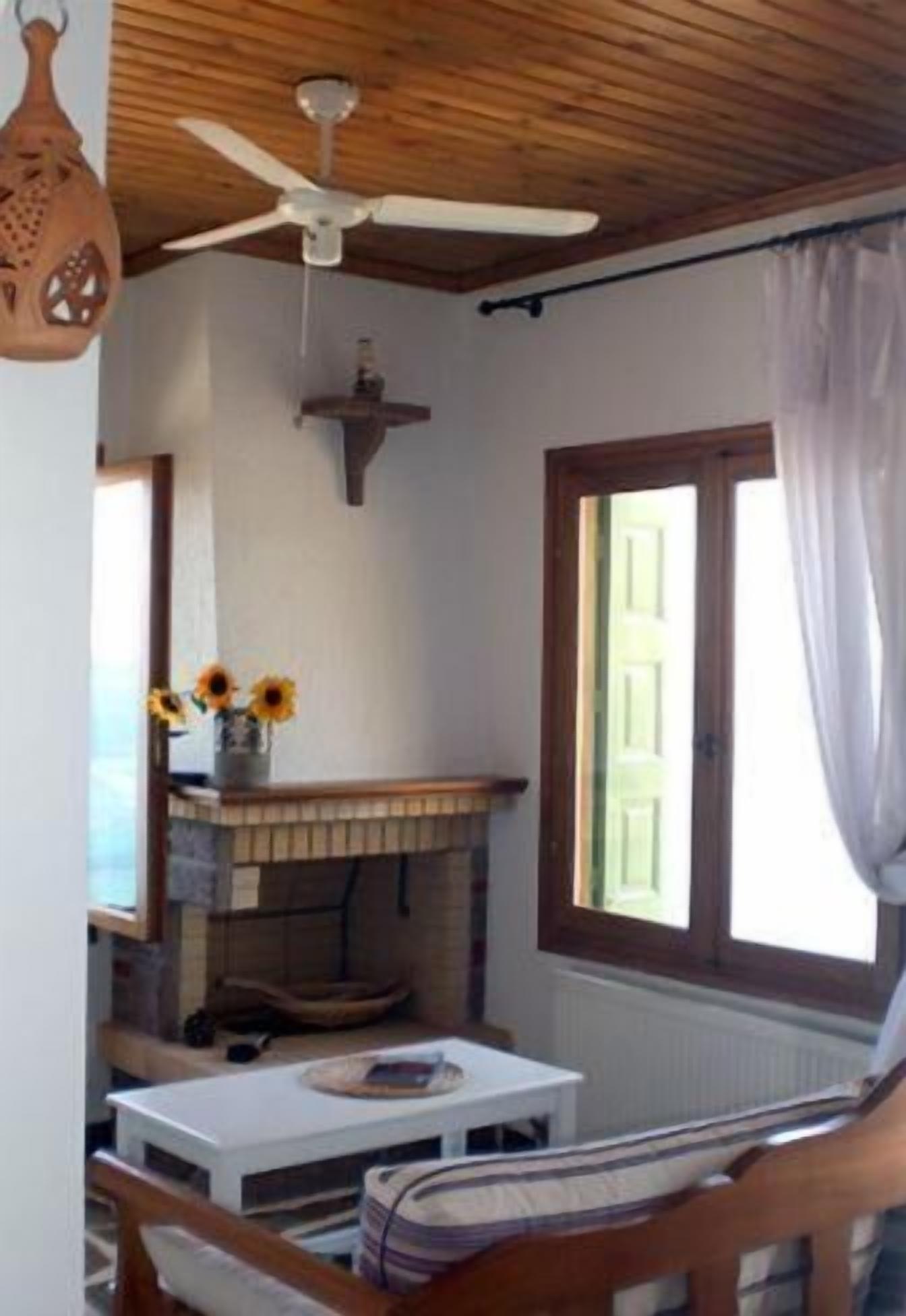 Una camera accogliente con soffitto in legno, una ventola a soffitto, un caminetto, una finestra con tende e un tavolo con un vaso di girasoli.
