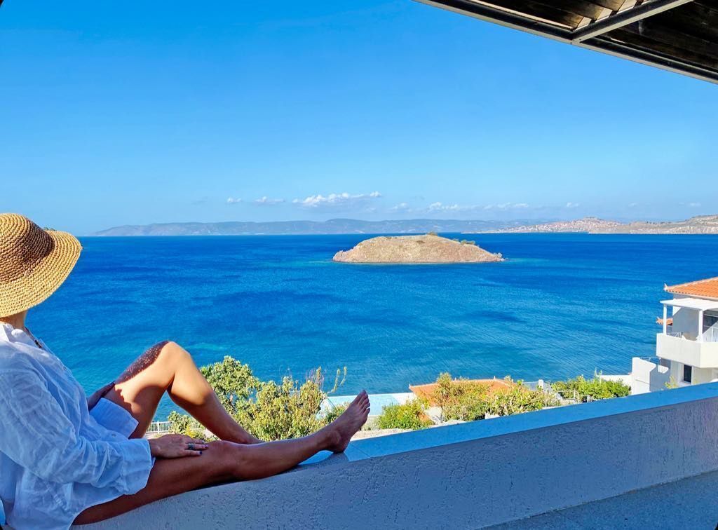 Una persona sentada en un balcón con vistas a un mar azul y despejado, con una pequeña isla en la distancia.