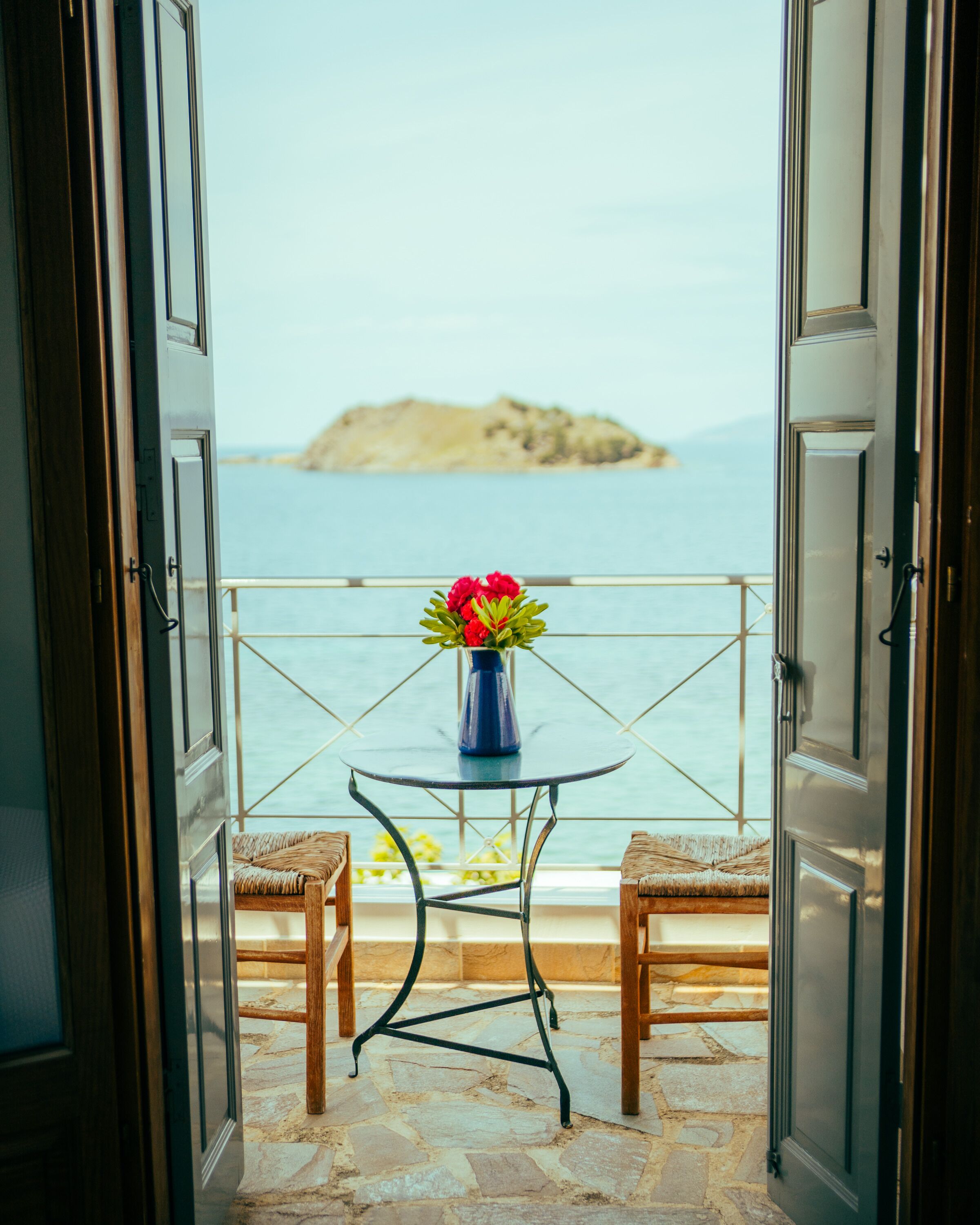 Een open balkon met een tafel en stoelen, een bloemenvaas en uitzicht op zee en een eiland in de verte.