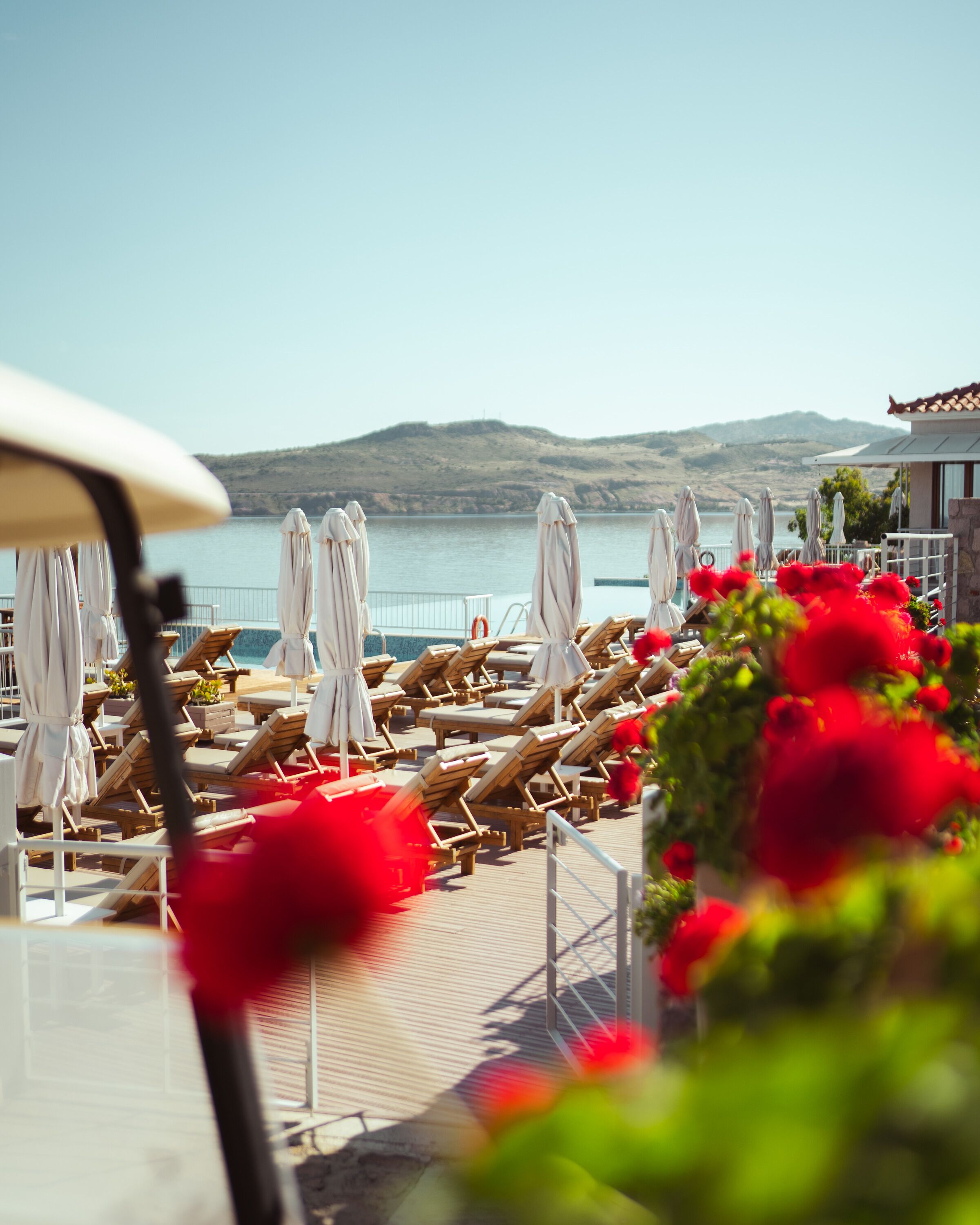 A beachside lounge area with sun loungers and umbrellas, overlooking a calm sea and distant hills.
