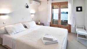 Une chambre avec un grand lit, une table de chevet ornée d’un vase de fleurs, une vue sur la mer par la fenêtre et une chaise blanche près de la fenêtre.