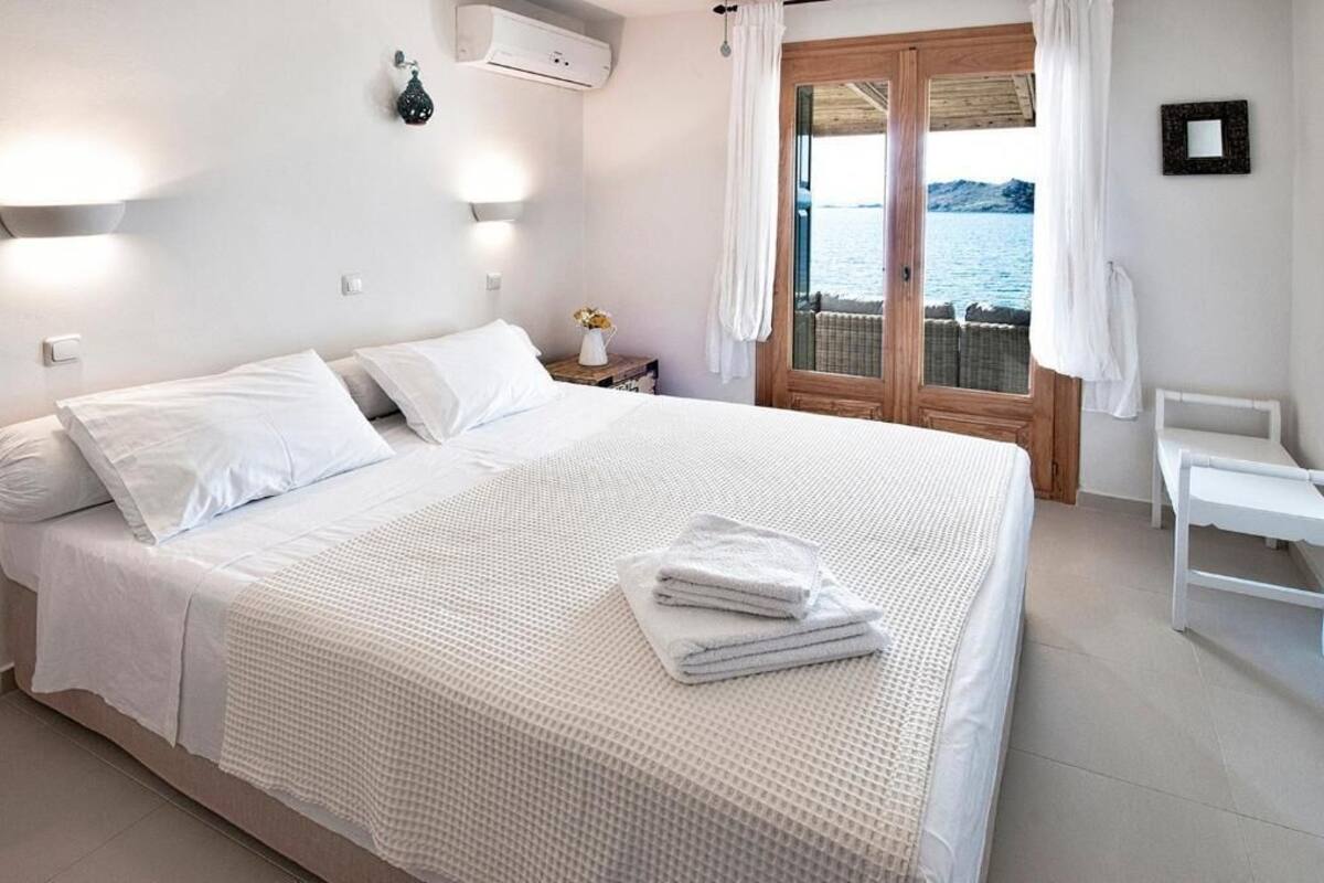 A bedroom with a large bed, a nightstand with a vase of flowers, a view of the sea through a window, and a white chair by the window.