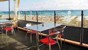 Een terras met tafels en stoelen, met uitzicht op een strand met parasols en ligbedden.