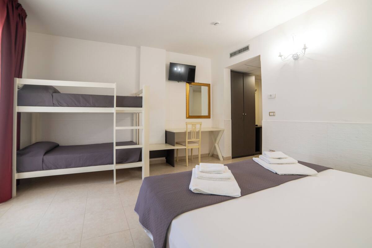 A hotel room with bunk beds, a desk, a chair, a TV, and a bed with towels.