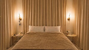 Een groot bed met een donkerbruine sprei, beige gordijnen en twee nachtkastjes met lampen.