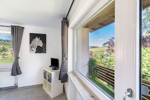 A balcony with a view of a green landscape, a white wall with a zebra painting, a small white shelf with books, and a black object on the shelf. - Little Cowhorse Ranch Apartment – Nature, Horses & Pure Tranquility (Münsingen)