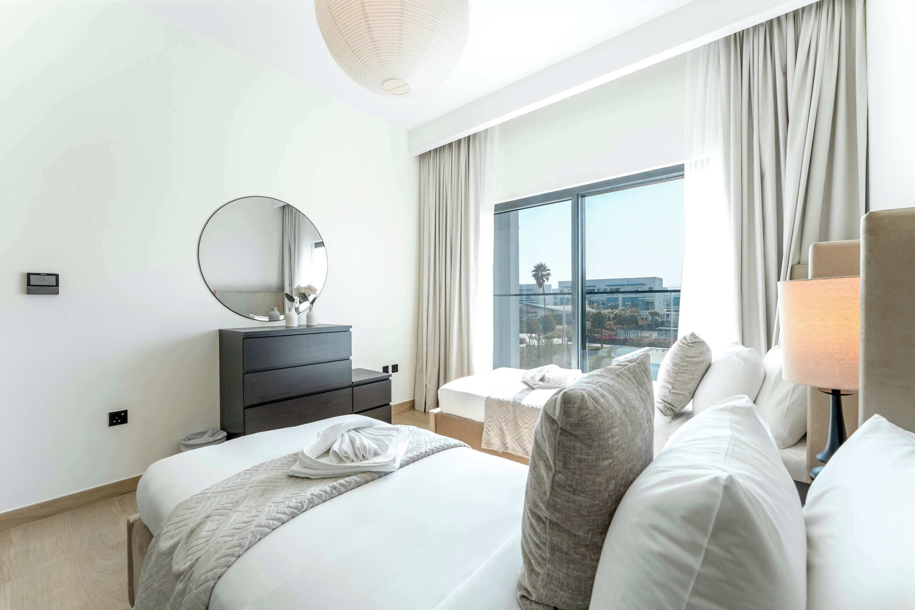 A modern hotel room with a large bed, a bedside table with a mirror, a small desk, and a view of the city through large windows.