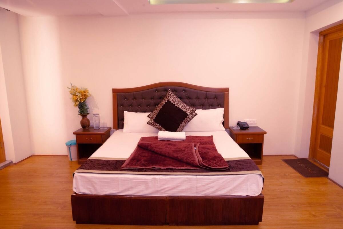 Een netjes opgemaakt bed met een bruin hoofdbord, witte lakens en een rode plaid. Er staan twee nachtkastjes bij, met lampen en een bloemenvaas op één.