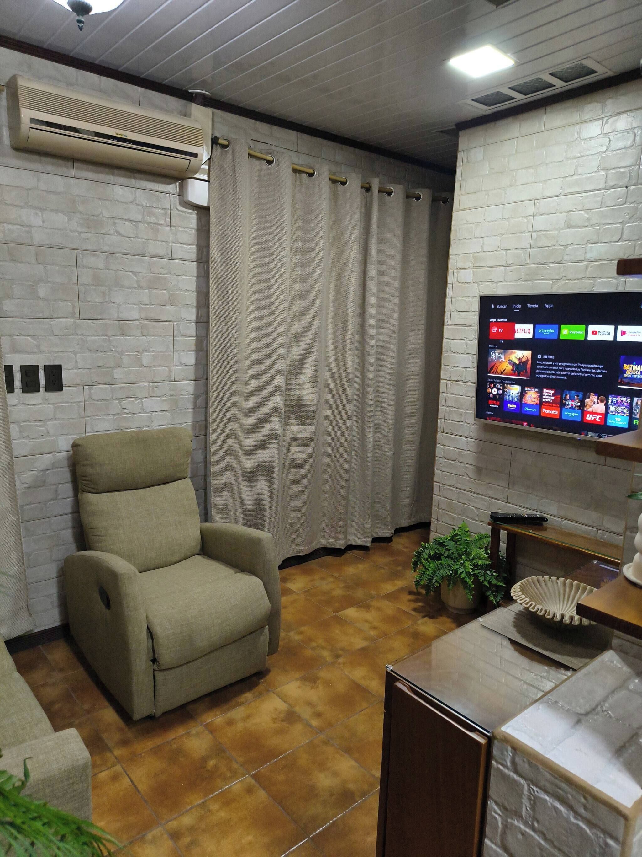 Una sala de estar con un sillón color beige, un televisor de pantalla plana montado en una pared de ladrillo y un ventilador de techo.