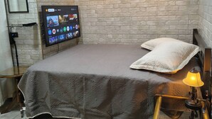 Makuuhuoneessa on tiilikuviollinen seinä, harmaalla päiväpeitteellä varustettu sänky, seinälle asennettu televisio, yöpöytä lampun kanssa ja tuoli.