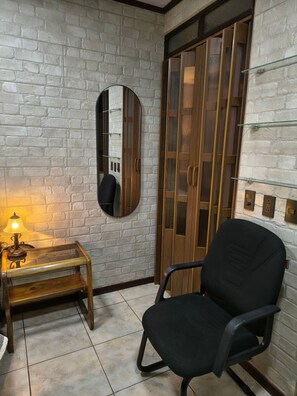 Una habitación con pared de ladrillo, un mueble de madera con puertas de cristal, una silla de oficina negra, una pequeña mesa de madera con una lámpara y un espejo en la pared.