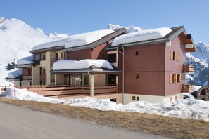 Un immeuble à plusieurs étages dont les toits sont recouverts de neige, avec un télésiège en arrière-plan et une chaîne de montagnes en toile de fond.