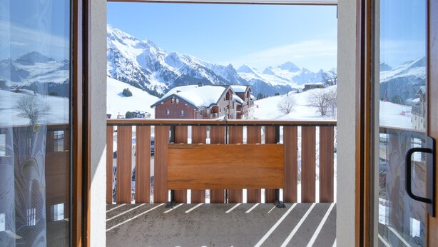 A balcony with a wooden bench, a view of snow-covered mountains, and a building in the background.