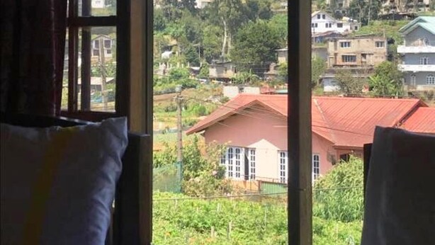 A room with a large window offering a view of a hillside with houses. There are two beds with white bedding. - Aurora Residence Nuwara (Nuwara Eliya)