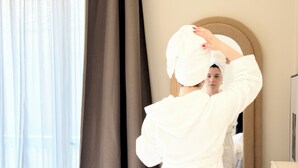 Ένα άτομο ντυμένο με μια λευκή ρόμπα, το οποίο διορθώνει μια πετσέτα στο κεφάλι του, βρίσκεται σε ένα δωμάτιο με έναν μεγάλο καθρέφτη, ένα παράθυρο με κουρτίνες και έναν μπεζ τοίχο.