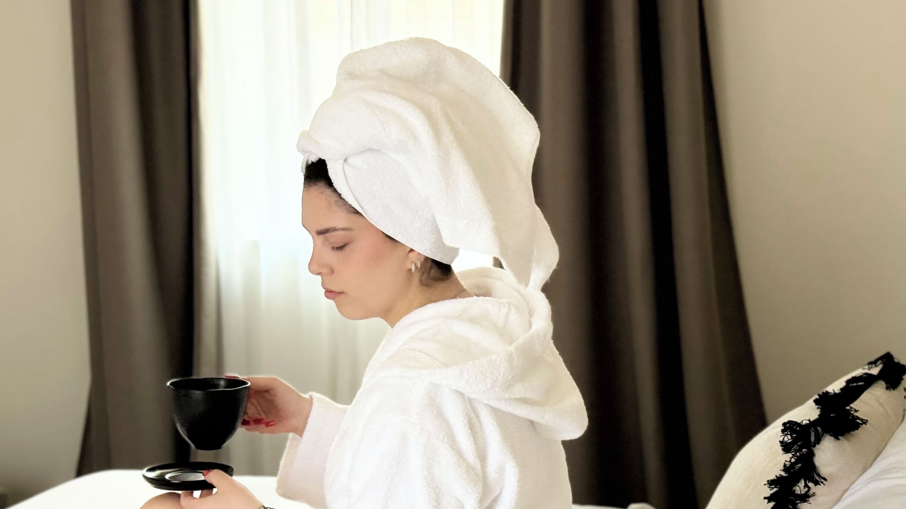 A person in a white robe with a towel on their head, holding a black cup, in a room with a bed and dark curtains.