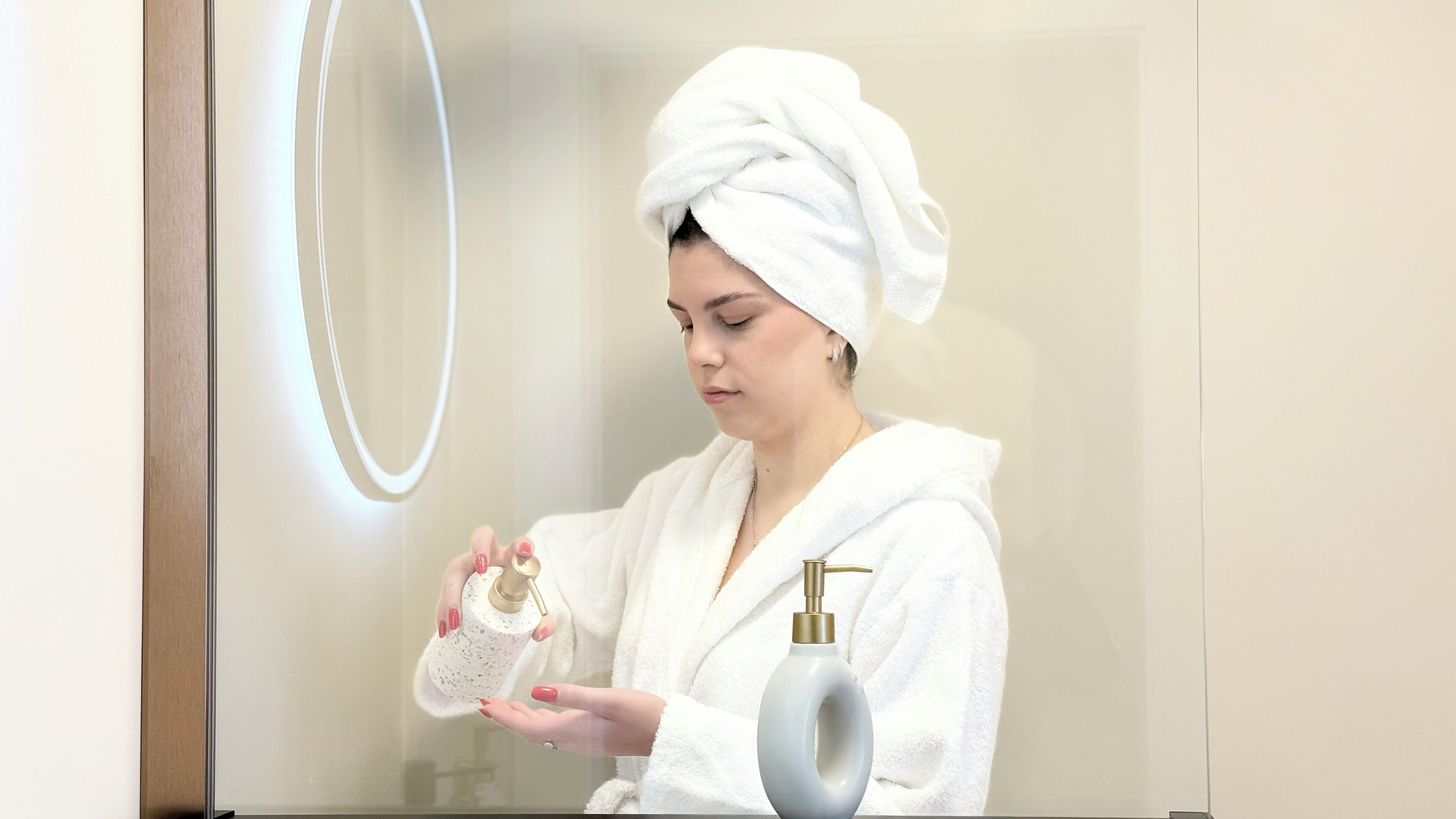 A person in a white robe holding a bottle of lotion and a pump bottle, standing in front of a mirror with a light ring.