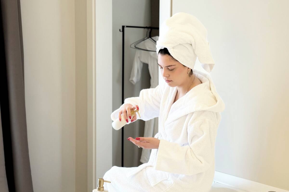 A person in a white bathrobe applying lotion in a bathroom with a towel rack and a white towel hanging on it.