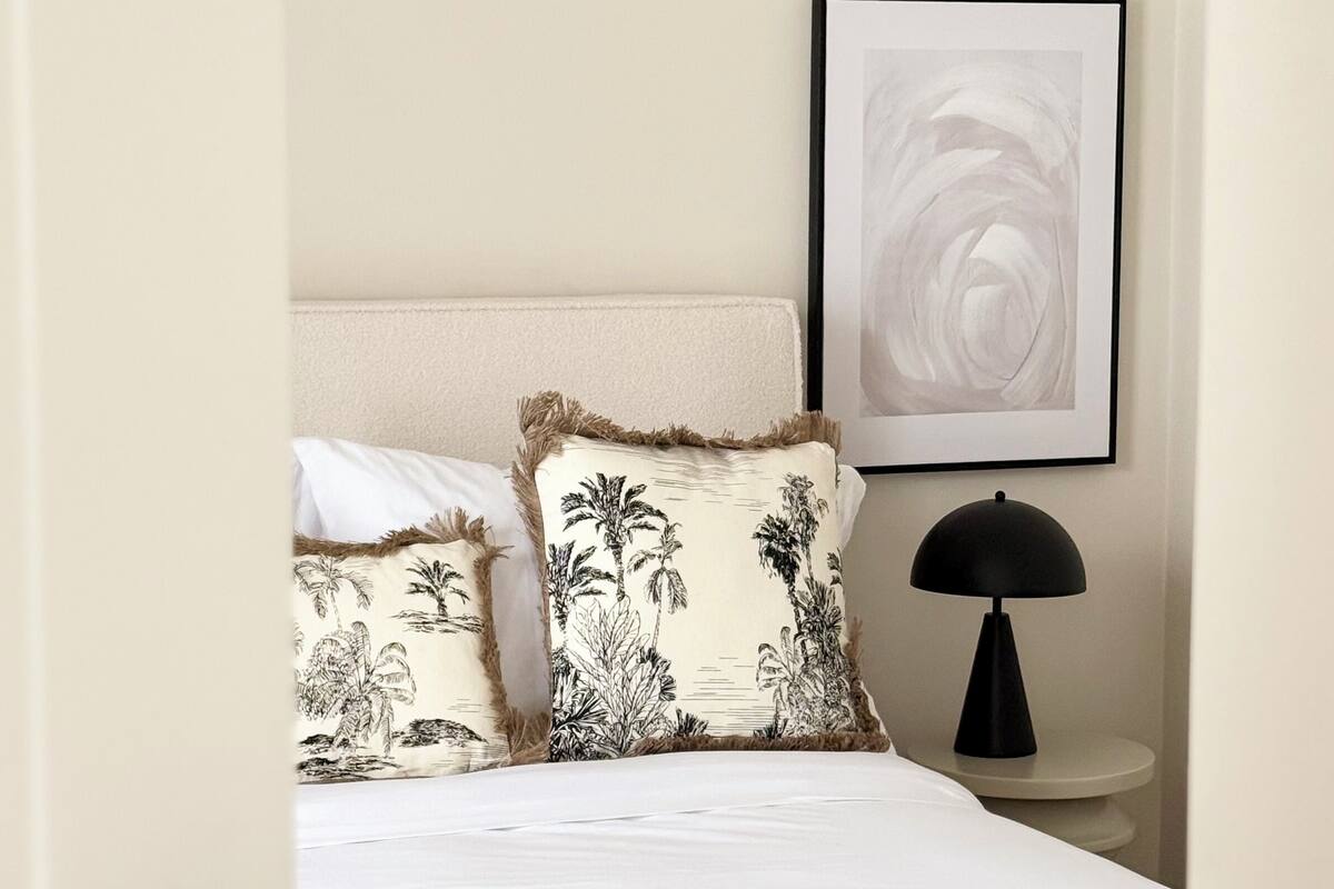 A bedroom with a bed, two pillows with tropical designs, a bedside table with a lamp, and a framed abstract painting on the wall.
