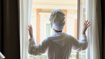 Una persona con bata blanca de pie junto a una ventana con cortinas, mirando hacia el exterior.