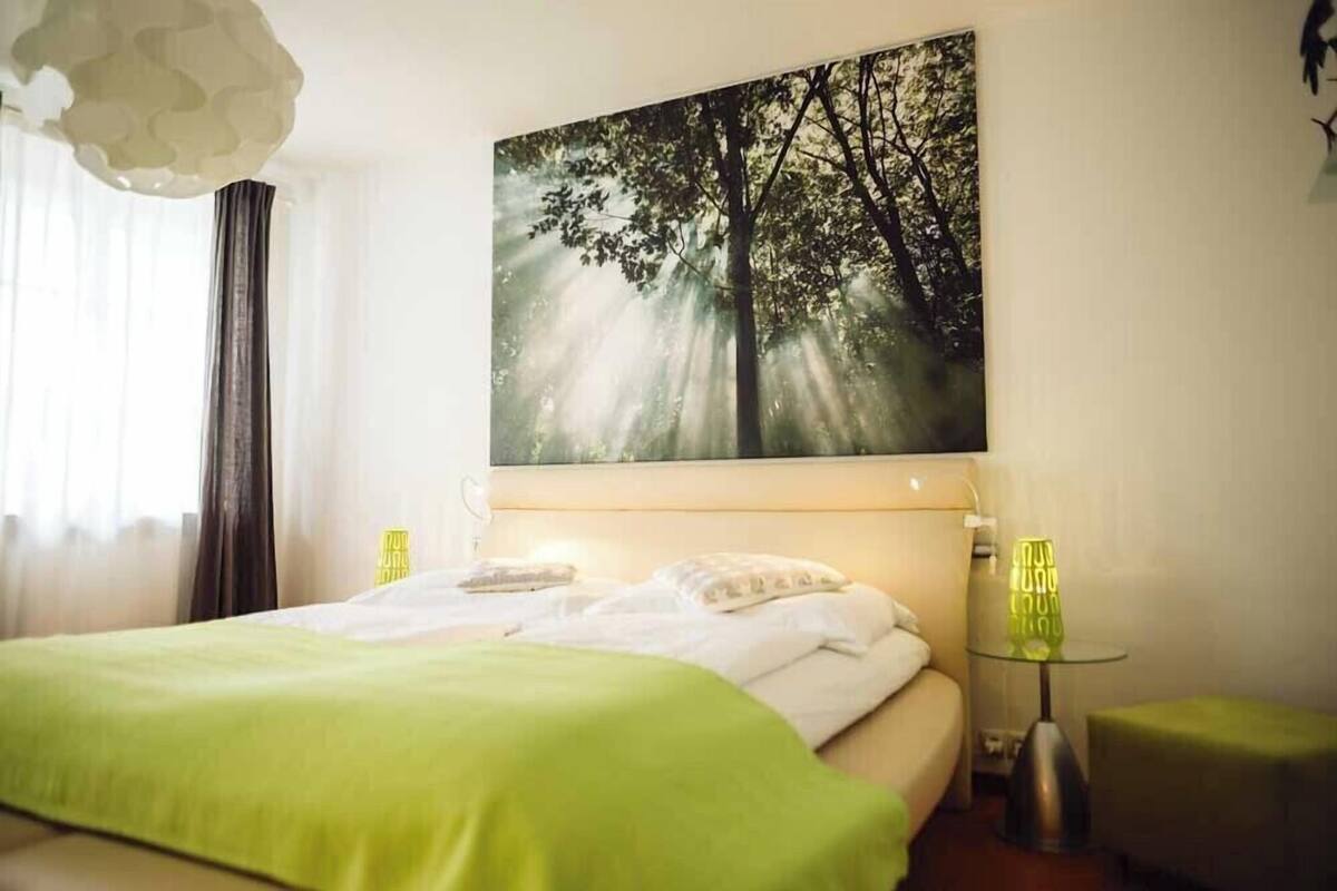 A bedroom with a bed, a green blanket, a wall-mounted painting of a tree, and a bedside table with a lamp.
