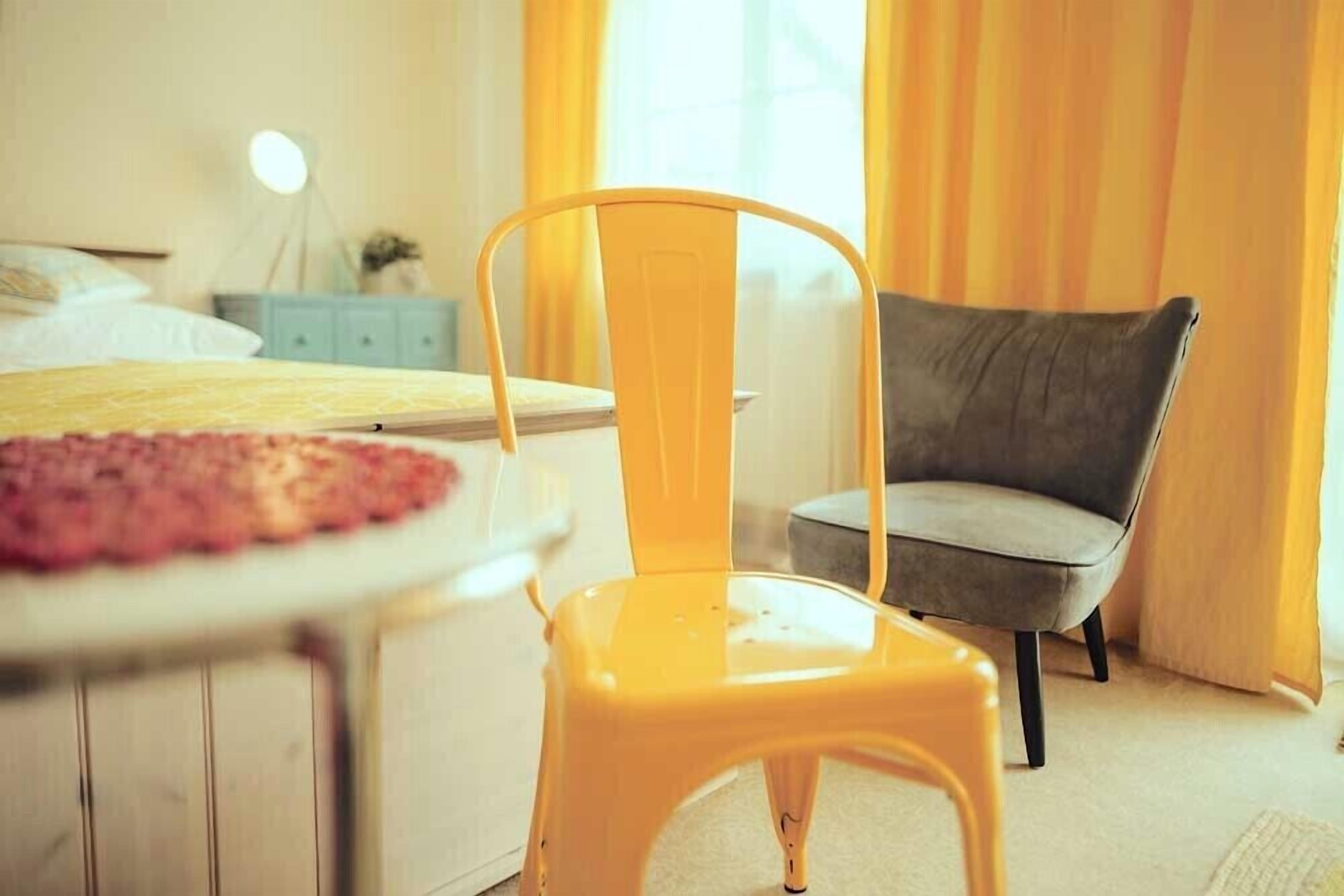 A bedroom with a bed, a yellow chair, a grey armchair, and a bedside table with a lamp.