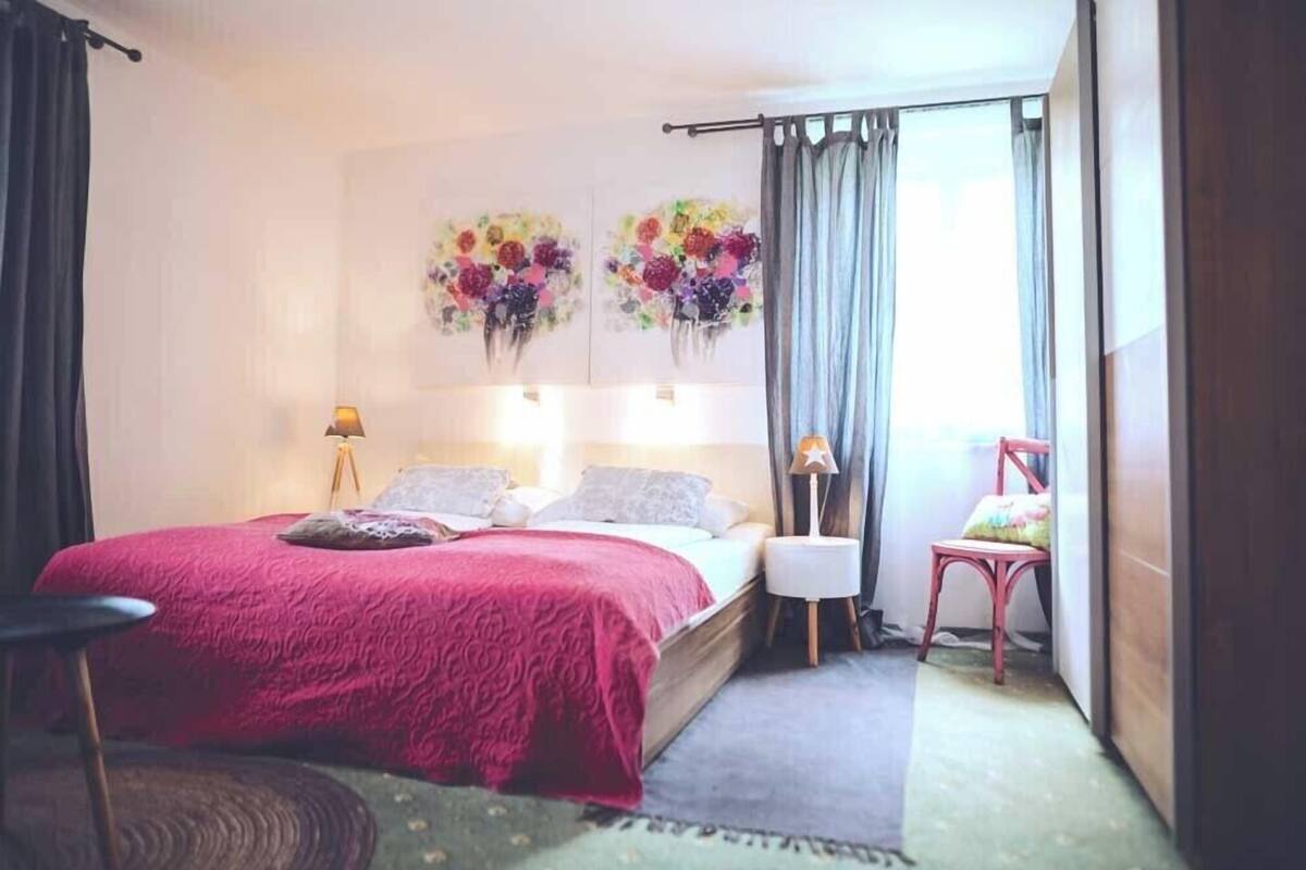 A bedroom with a bed, two wall-mounted lamps, a chair, and a window with curtains.