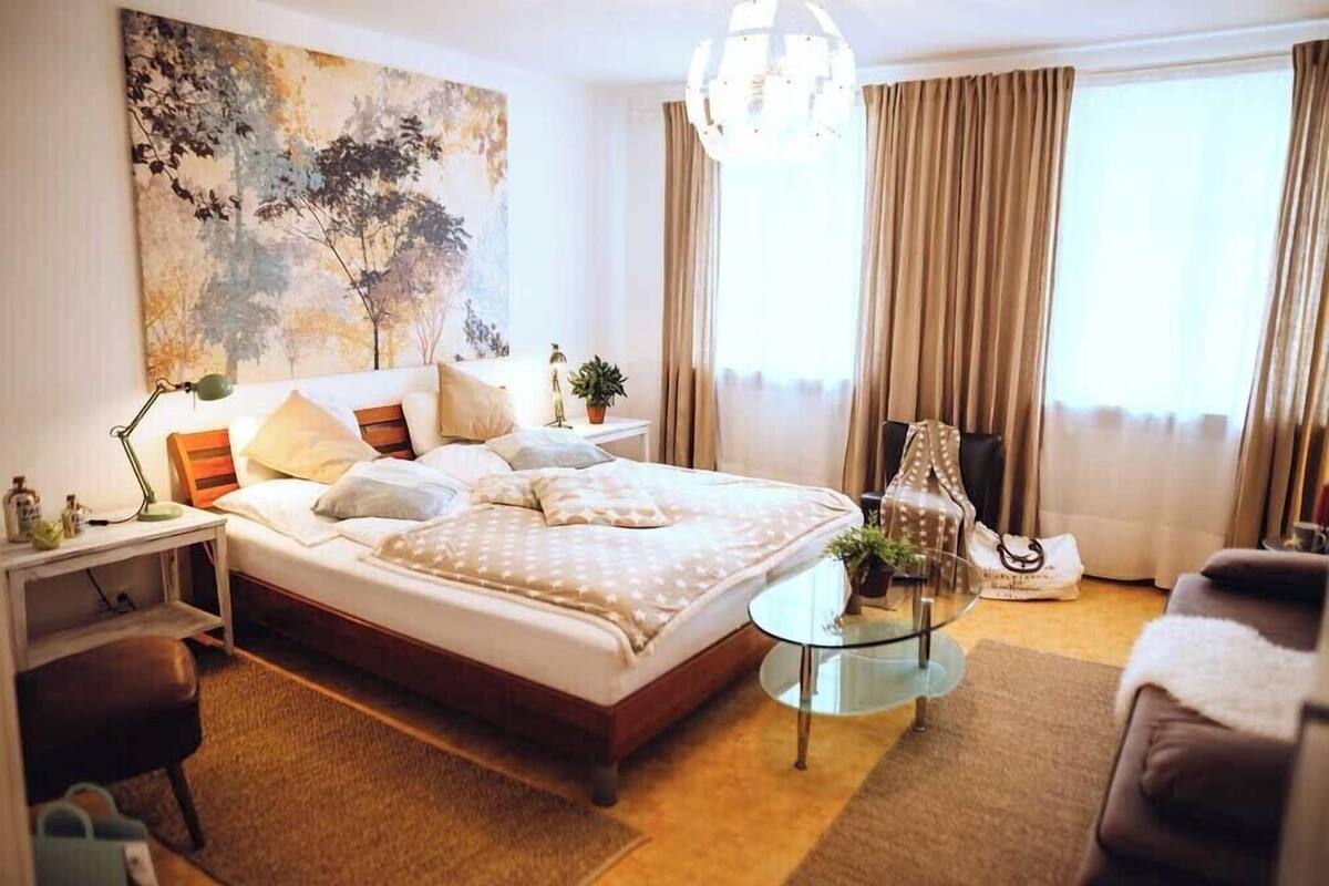 A well-lit bedroom with a large bed, a bedside table, a small round table, a chair, and a large window with curtains.