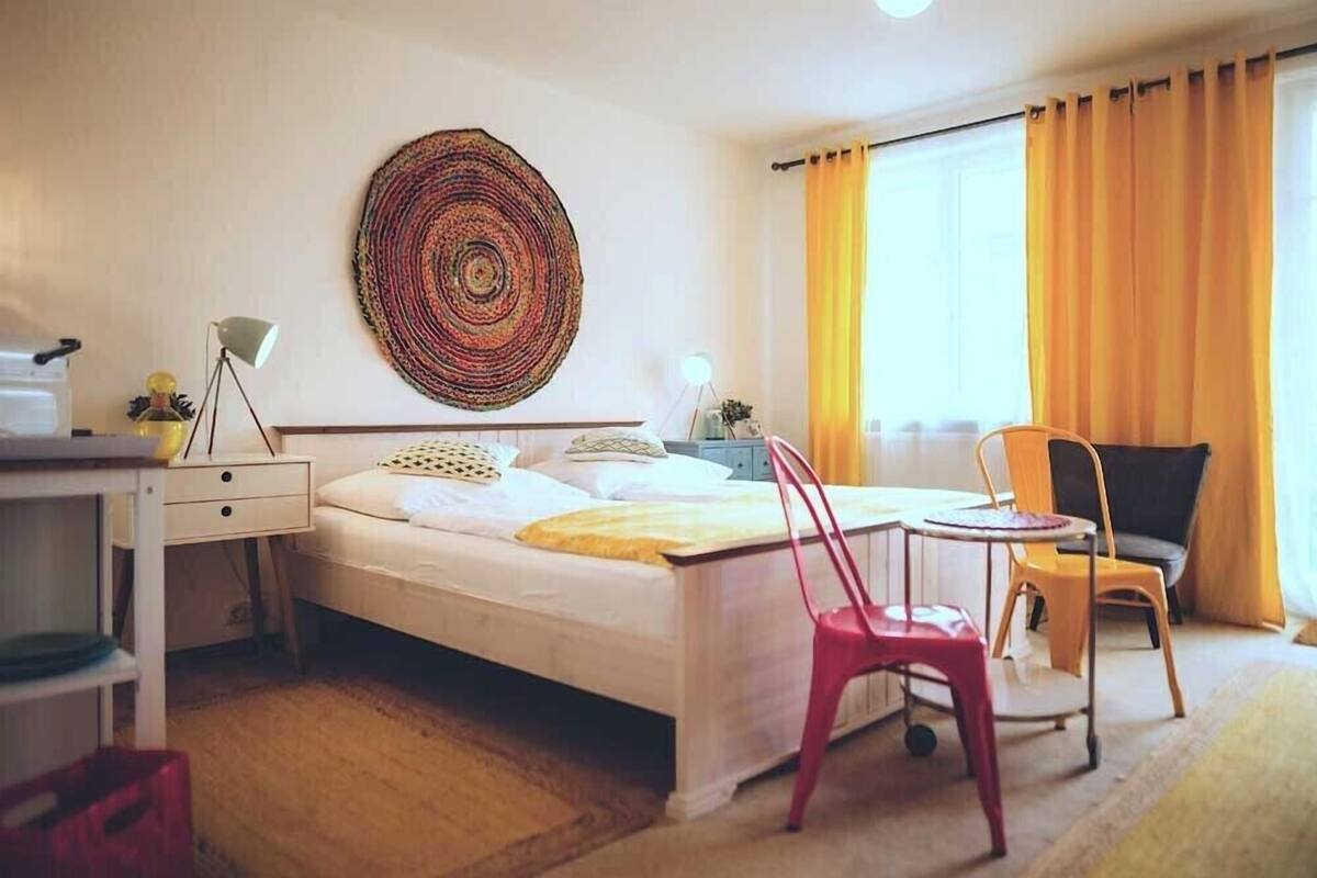 A hotel room with a bed, a desk, a chair, a lamp, and a large woven tapestry on the wall.