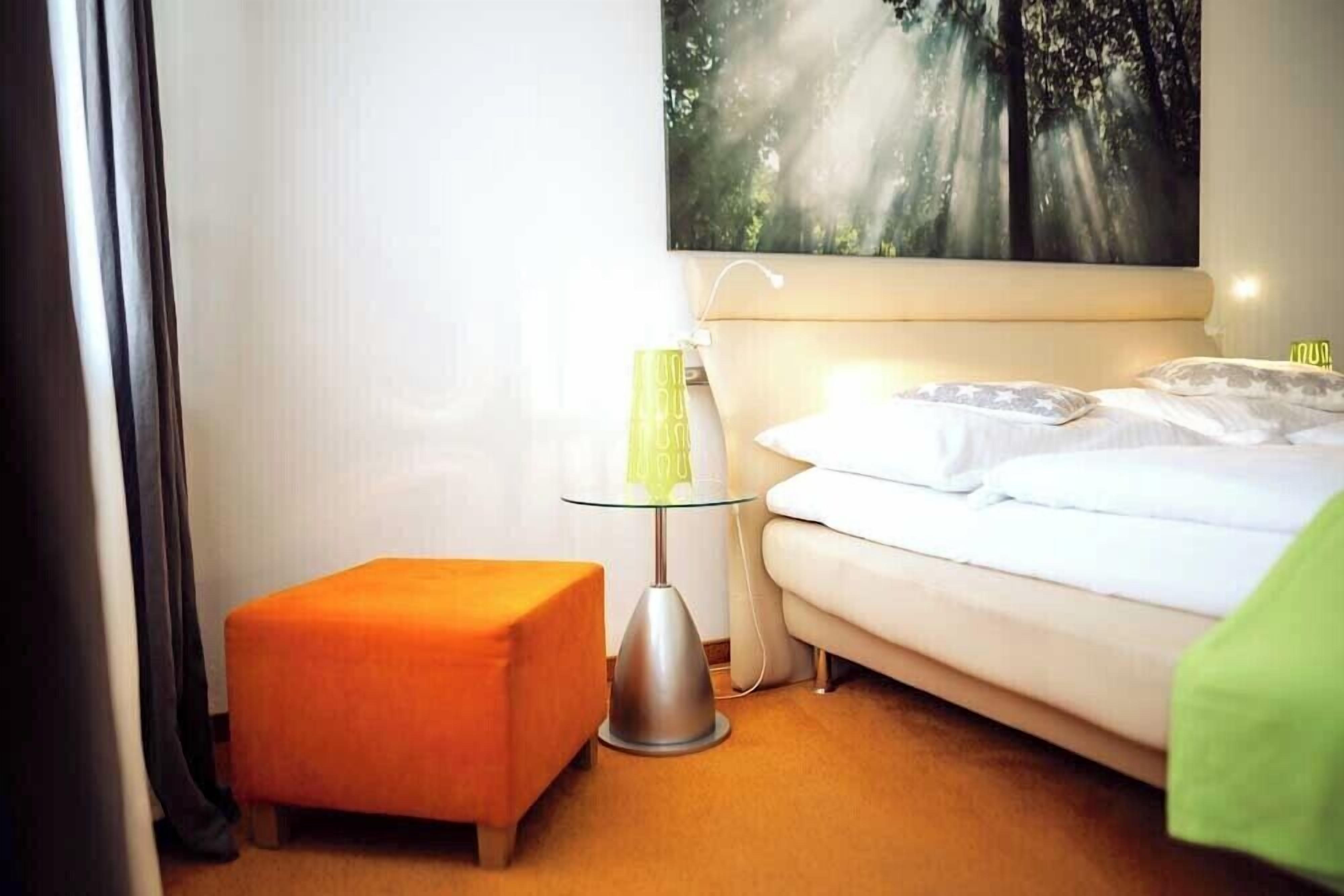 A hotel room with a bed, a small orange ottoman, a glass table with a green vase, and a painting of a forest on the wall.
