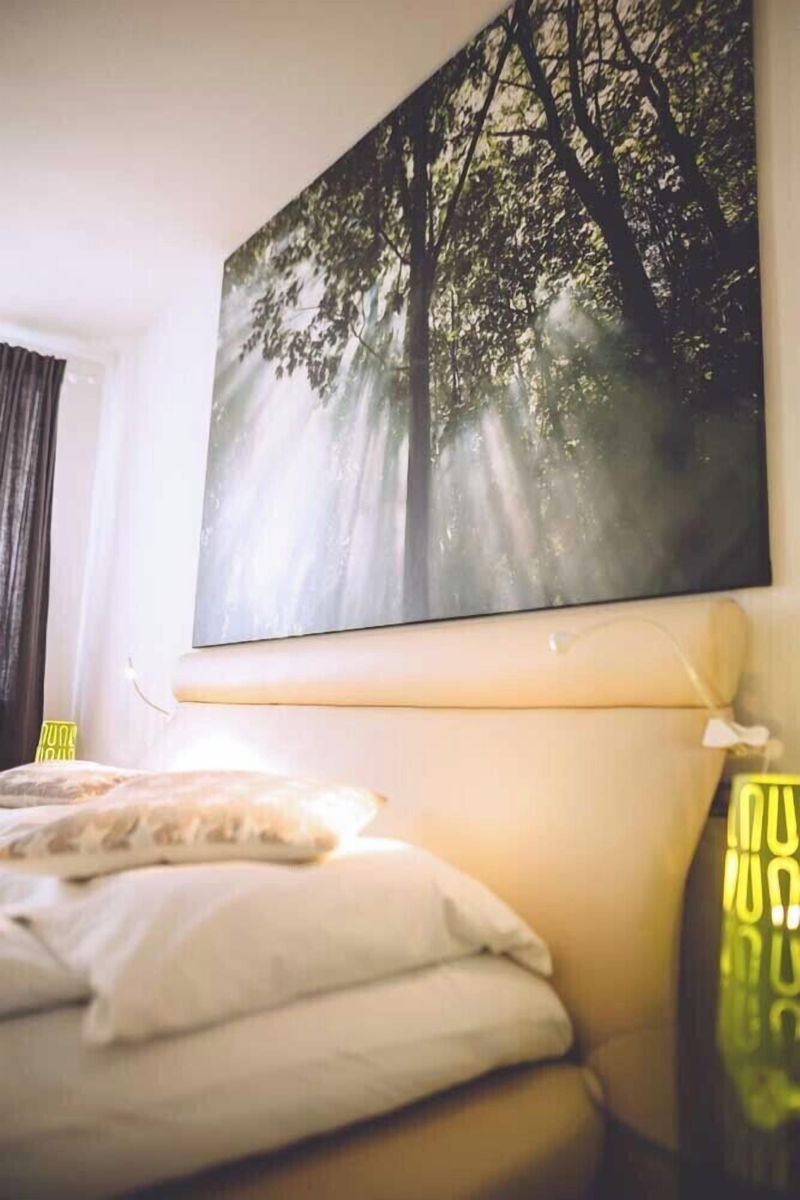 A bedroom with a bed, a bedside table, a lamp, and a large forest-themed painting on the wall.