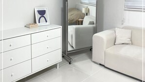 Une chambre moderne avec une commode blanche, une armoire à miroir, un canapé blanc et une fenêtre avec des stores.