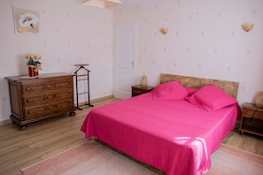 Une chambre à coucher avec une commode en bois, un lit recouvert d’une couvre-lit rose, une table de chevet en bois et une lampe fixée au mur.