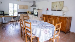 Un comedor con una mesa dispuesta para seis personas, sillas de madera, una cocina al fondo y un cuadro en la pared.